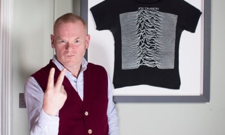 A portrait photo of George Shaw in a sleeveless marron cardigan and shirt, gesturing a V sign while looking directly at the camera in front of a poster featuring the cover of Joy Division's 'Unknown Pleasures' album cover