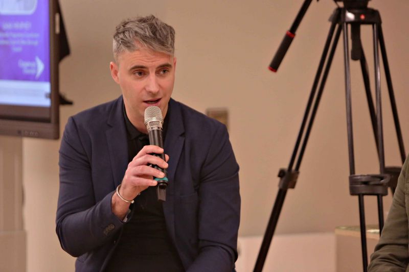 A man dressed in a formal suit holds a microphone, indicating his readiness to engage with listeners.