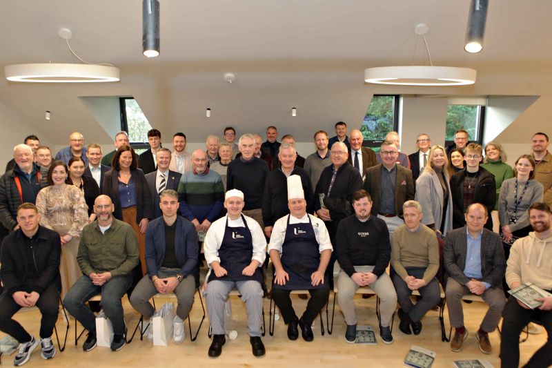 A group of people, some seated and some standing, pose for a photo in a well-lit room with large windows. Two chefs in uniforms are seated in the front row.