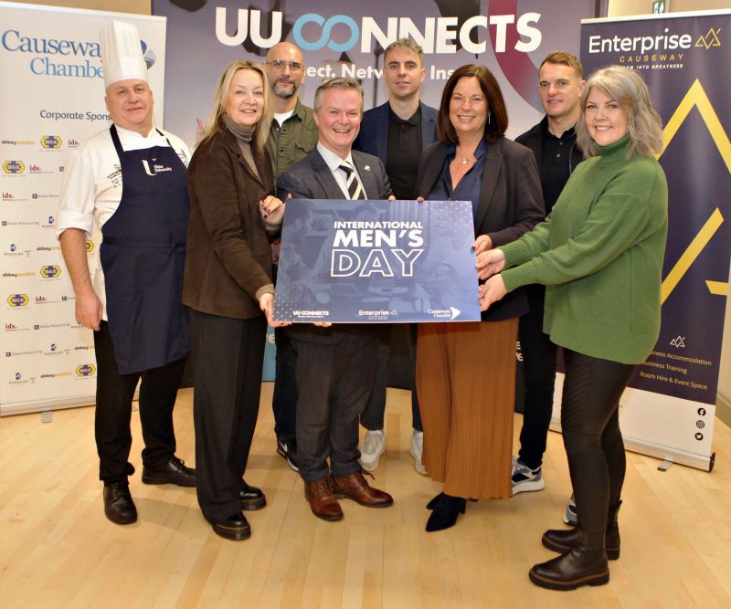 A group of eight people stands together indoors, holding a large sign that reads \