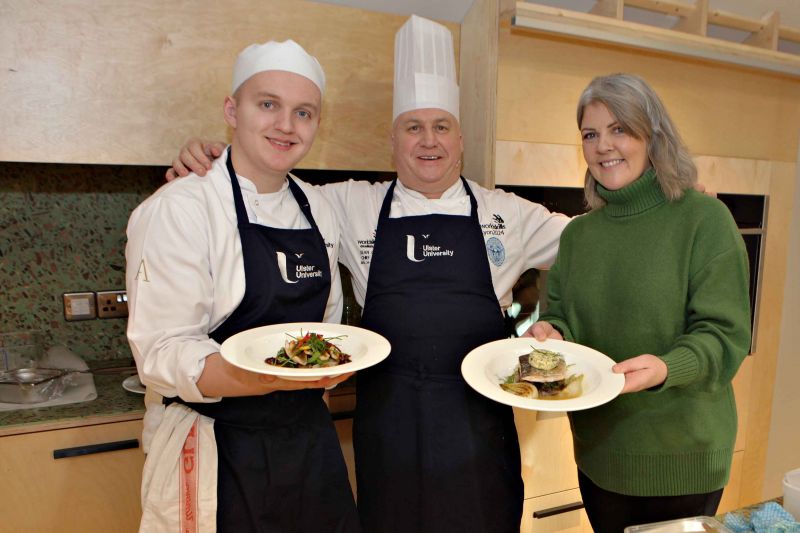 Two Chefs standing on the left hand side and in the middle standing beside a woman in green on the right hand side while showcasing the food they created for the International Men\'s Day Workshop 