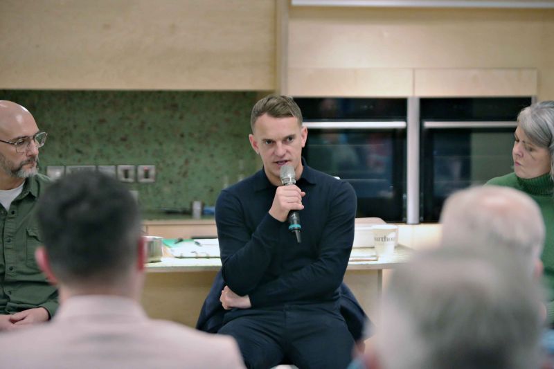 A man engages with an audience while speaking into a microphone, seated comfortably in a well-lit room 